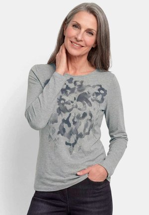 Smiling middle-aged woman with gray hair wearing a gray long-sleeve shirt with a dark floral pattern and dark pants, one hand on neck, the other in pocket.