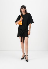 Black t-shirt dress with a knotted front detail, short sleeves, and a small logo. Paired with a bright orange mini bag and black heels.