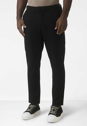 Mustad vöötrükiga püksid kerge kangaga, kohandatud lõikega, küljetaskutega ja mustade tennistega.