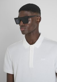 Gafas de sol negras de gran tamaño con forma angular, combinadas con una camiseta polo blanca con cuello acanalado y un logo en relieve en el pecho.