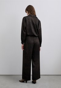 Tweedelig ensemble met een donkere, textuurrijke stof. Het bovenstuk heeft een kraag en lange mouwen; de broek heeft een wijde pijp en een hoge taille.