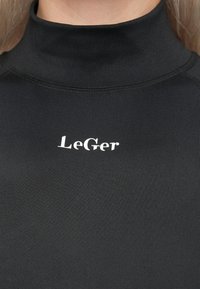 Svart høyhalset topp med hvit "LeGer" logo sentrert på brystet, båret av en person med lys hud og blonde hår.