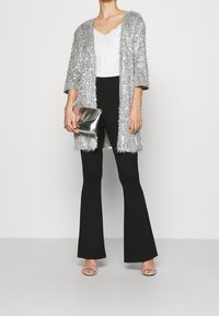 Silvrig paljettkimono över en vit topp, ihop med svarta utsvängda byxor och silvriga sandaler med remmar. Håller en metallisk kuvertväska.