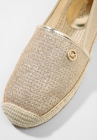 Alpargata de tono dorado con una parte superior texturizada, bordeada por un ribete metálico. Presenta una suela de yute tejido y un pequeño detalle con el logo.