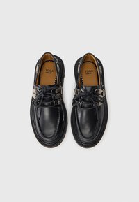 Chaussures en cuir noir avec un bout rond, des coutures détaillées et des accents métalliques uniques sur les côtés. Elles présentent un design à lacets et des semelles texturées.