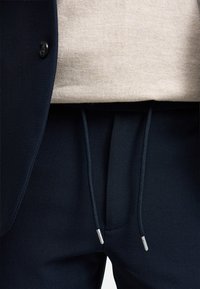 Pantaloni jogger blu navy con finitura testurizzata, dotati di vita con cordoncino e punte argentate, abbinati a un maglione beige chiaro sotto un blazer.