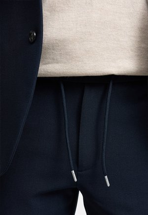 Marineblå joggebukser med struktureret finish, der har elastisk talje med snoretræk og sølvfarvede spidser, parret med en lys beige sweater under en blazer.