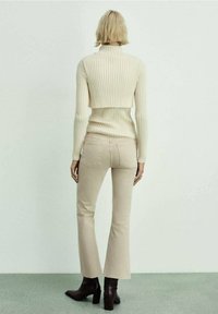 Femme aux cheveux blonds courts portant un col roulé court côtelé couleur crème, un pantalon beige ajusté et des bottes à talons marron foncé, se tenant face à un mur blanc.