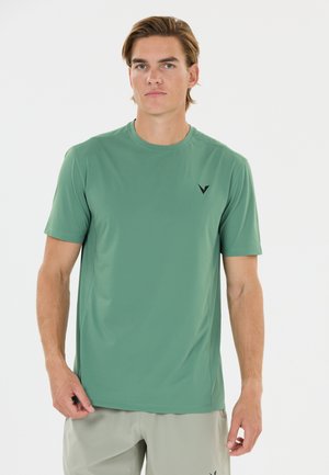 Junger Mann mit hellbraunen Haaren, der ein grünes kurzärmeliges T-Shirt und hellgraue Shorts trägt, steht vor einem weißen Hintergrund.