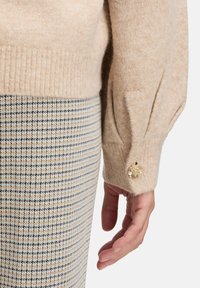Arm in einem beigen Strickpullover mit Puffärmel und goldenen Blumenknopf, der neben einem Rock mit beige, schwarz und weißem karierten Muster ruht.