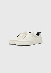 White sneakers with a suede toe cap, textured side panels, and black accents. Features flat laces and a thick rubber sole.