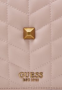 Prošívaný růžový syntetický materiál se zlatým pyramidovým cvokem a vyraženým logem "GUESS EST 1981" ve spodní části, s kontrastním prošitím.