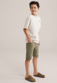 Kratki rukavi bež T-shirt s malim logom, uparen s maslinasto zelenim kratkim hlačama do koljena i smeškim nubuk cipelama na slip.