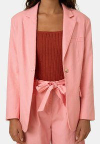 Helles rosa Blazer mit einem einzelnen Knopf und Reverskragen, kombiniert mit einem strukturierten rostfarbenen Oberteil und passenden hochtaillierten Hosen.