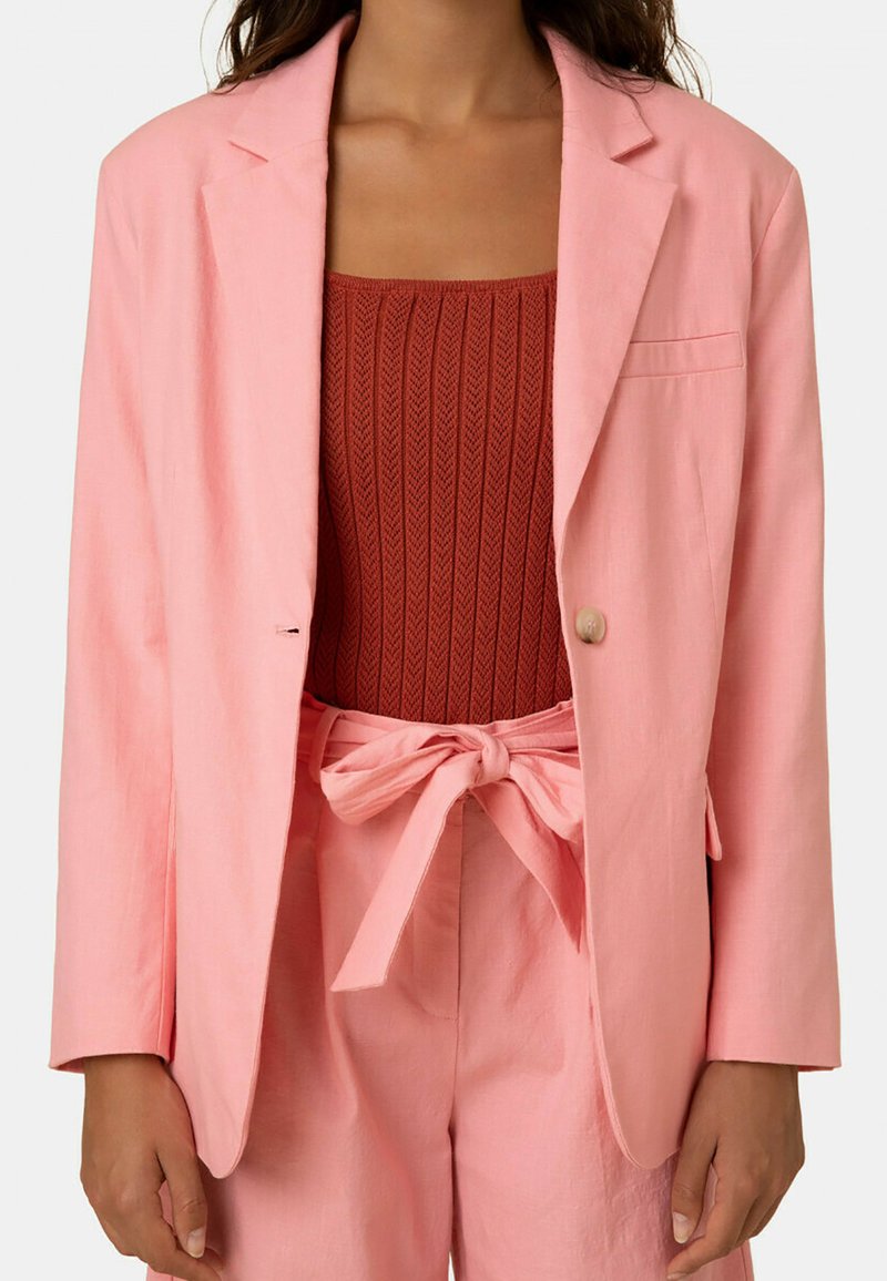 Helles rosa Blazer mit einem einzelnen Knopf und Reverskragen, kombiniert mit einem strukturierten rostfarbenen Oberteil und passenden hochtaillierten Hosen.