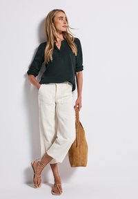Donna che indossa una maglietta a maniche lunghe verde scuro, pantaloni bianchi a gamba larga e cropped, sandali beige e porta una grande borsa in suede color nocciola.