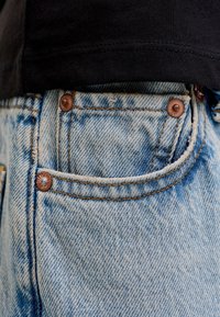 Záber na predné vrecko svetlomodrých riflí z denimového materiálu s medenými nitmi, nosené s čiernou košeľou.