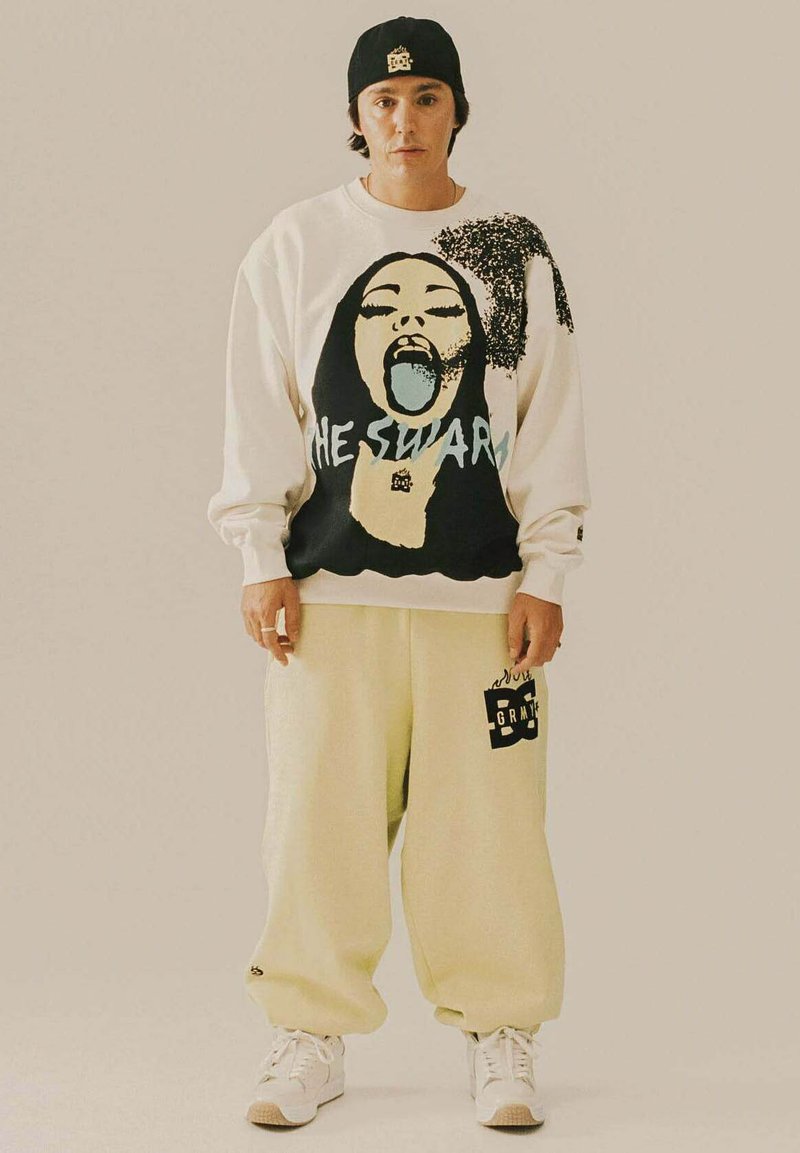 Persoon staat in een zwarte pet, witte sweatshirt met een grafisch gezicht en tekst, crèmekleurige baggy broek en witte sneakers.