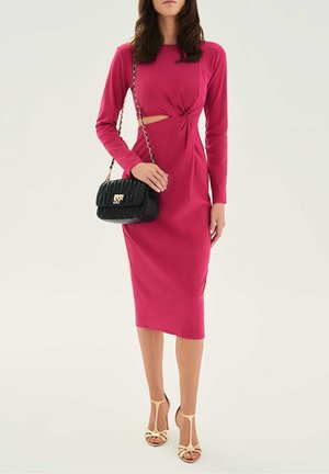 Fuchsia lange mouwen jurk met een gedraaide detail en een uitsnijding bij de taille, gecombineerd met een gewatteerde zwarte handtas en naaktkleurige sandalen met hak.