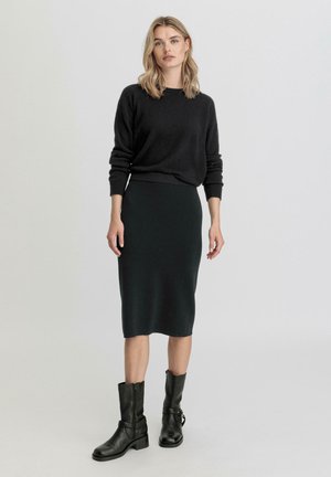Maglione nero, gonna verde scuro fino al ginocchio e stivaletti neri in pelle con punta quadrata. La modella è in piedi con le mani sui fianchi.