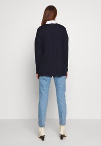 Haut à manches longues marine avec un col rond et des fentes latérales, associé à un jean bleu clair et des bottines crème, sur fond blanc.