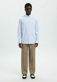 Camisa a rayas azul claro con diseño de botones, mangas largas y cuello clásico; combinada con pantalones de pierna ancha beige y zapatos bicolor.