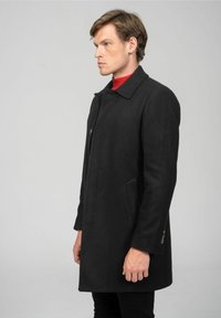 Zwart wollen jas met een kraag, rechte snit, zijzakken en knoopsluiting. Gedragen over een rode coltrui, weergegeven tegen een grijze achtergrond.