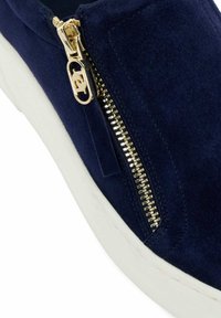 Baskets en suède bleu marine avec une semelle blanche et un détail de fermeture éclair doré. La fermeture éclair est dotée d'une tirette circulaire pour une ouverture et une fermeture faciles.