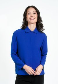 Kék, hosszú ujjú blúz gallérral és elülső cipzárral, sima textúrával és laza szabással, fekete nadrággal viselve.