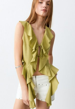 Jeune femme portant un chemisier sans manches vert olive à volants noué devant, short blanc taille haute et un bracelet argenté au bras.