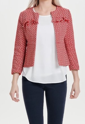 Vrouw draagt een rood gestructureerd cropped jasje over een witte blouse en donkere aangesloten broek tegen een effen achtergrond.