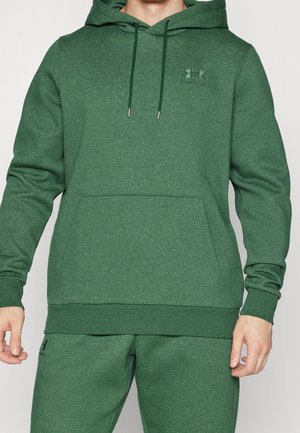 Homme portant un sweat à capuche vert Under Armour avec poche avant et un pantalon de survêtement vert assorti sur un fond uni.