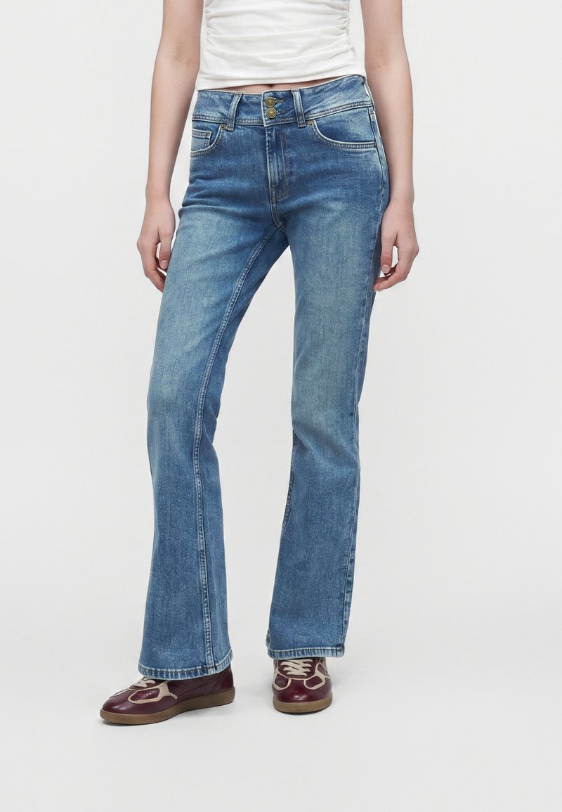Άτομο που φοράει μεσαίου μπλε τζιν bootcut, λευκή κοντή μπλούζα και μπορντό και μπεζ αθλητικά παπούτσια, στέκεται σε λευκό φόντο.