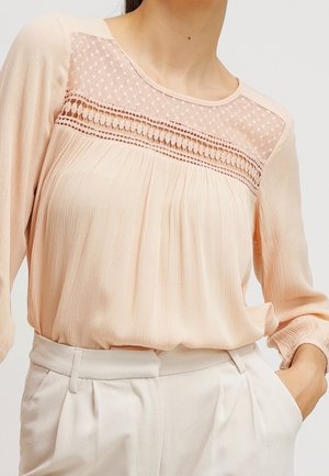 Blusa lunga color pesca chiaro con yoke in pizzo testurizzato, davanti plissettato e vestibilità rilassata. Indossata con pantaloni a vita alta di colore chiaro.