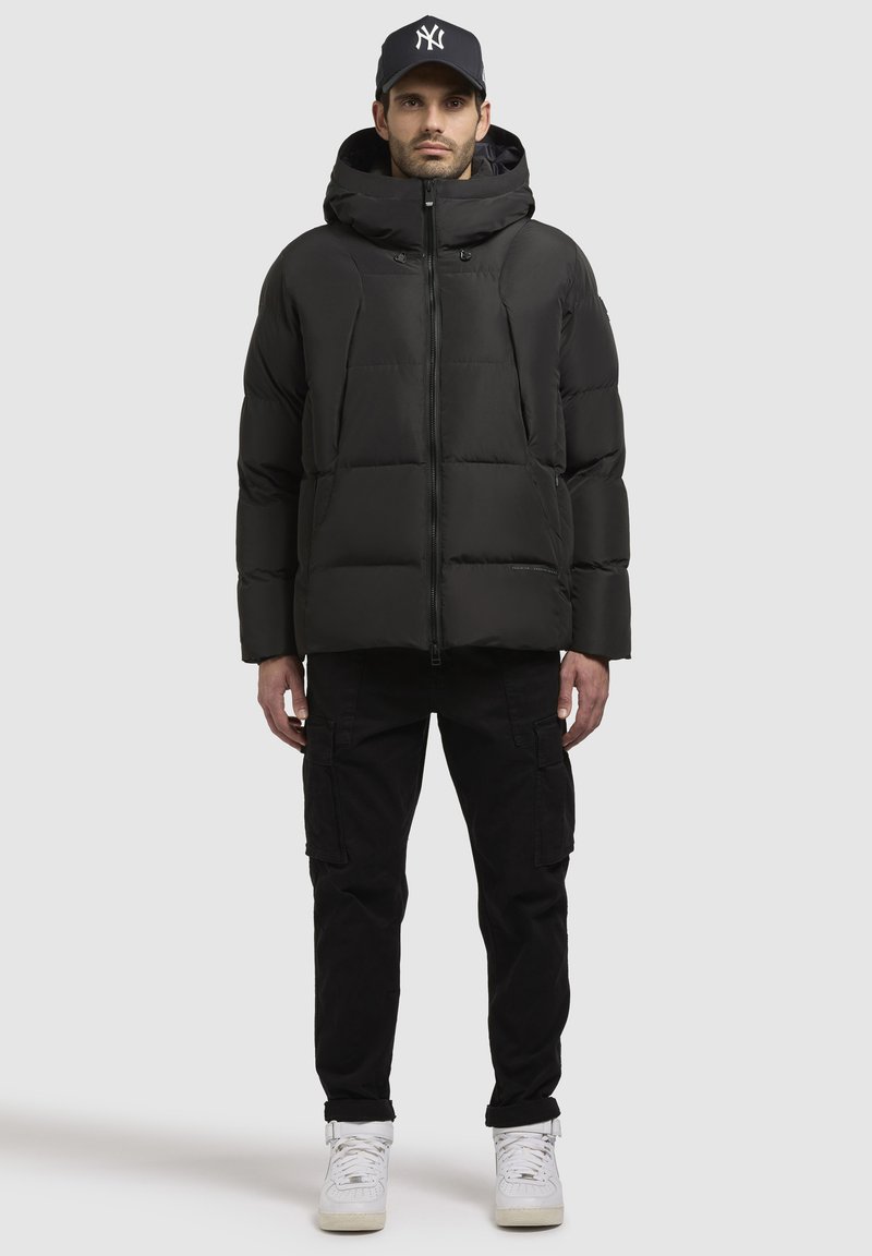 Schwarze Pufferjacke mit Kapuze, ausgestattet mit einem Reißverschluss und sichtbaren Nähten. Kombiniert mit schwarzen Cargo-Hosen und weißen High-Top-Sneakern.