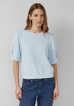 Frau trägt ein hellblaues Kurzarmshirt mit Puffärmeln und blaue Jeans, steht vor einem schlichten hellgrauen Hintergrund.