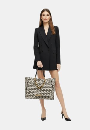 Donna con abito blazer doppiopetto nero che tiene una grande borsa tote beige e nera con manici a catena dorata, indossando tacchi alti neri.