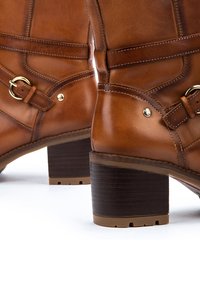 Des bottines en cuir marron possèdent un bout arrondi, des accents en quincaillerie dorée, et un talon empilé avec des détails texturés sur la semelle.