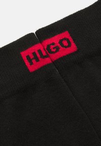 Calcetines negros con textura acanalada, con una banda roja en la parte superior que lleva la inscripción "HUGO" en letras negras. Diseño sencillo, sin patrones adicionales.