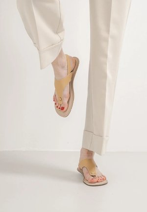 Pies con sandalias beige de punta abierta con correas en el tobillo y esmalte de uñas rojo, vistiendo pantalones color crema con puños anchos.