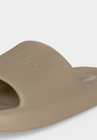 Μπεζ σανδάλι slip-on με φαρδιά λουράκι που φέρει ανάγλυφη επιγραφή "GUESS" στο πάνω μέρος.