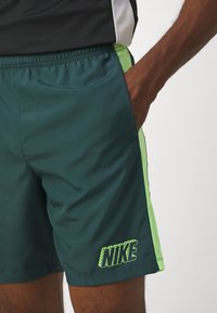 La mano de un hombre en el bolsillo de unos pantalones cortos Nike verdes oscuros con franjas laterales en verde neón, vistiendo una camiseta en blanco y negro.