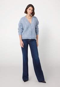 Hellblauer, gestrickter Cardigan mit V-Ausschnitt, strukturiertem Rautenmuster und Knopfverschluss, kombiniert mit dunkelblauen Schlagjeans und schwarzen High Heels.