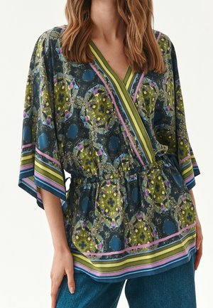 Frau trägt eine farbenfrohe, floral gemusterte Wickelbluse mit weiten Ärmeln und gestreiftem Besatz über blauen Jeans.