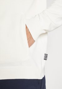 Suéter branco de malha com punhos e bainha canelados, apresentando um bolso lateral e uma pequena etiqueta de marca na borda inferior. Textura suave.
