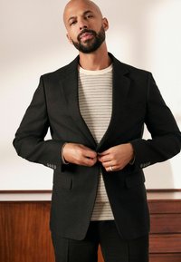 Blazer negro con solapas de muesca y dos bolsillos frontales, superpuesto a una camiseta a rayas beige y blanca. Modelo ajustando los botones del blazer.