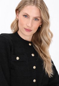 Černý cardigan s zlatými knoflíky a malou náprsní kapsou, vyrobený z měkké texturované látky s kulatým výstřihem.