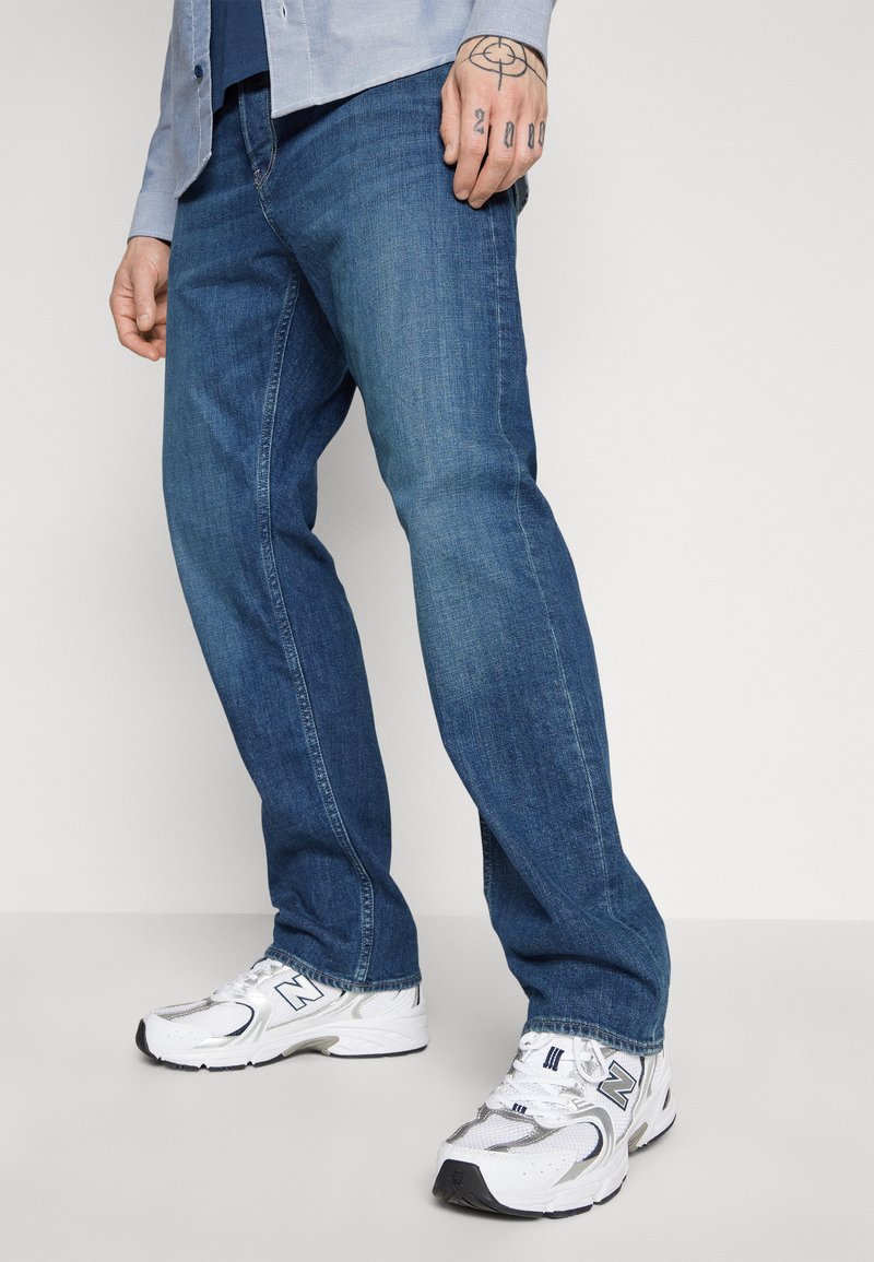 Jeans en denim bleu avec une coupe droite, présentant un léger délavage et des coutures contrastées. Assortis à des baskets blanches avec des accents gris.