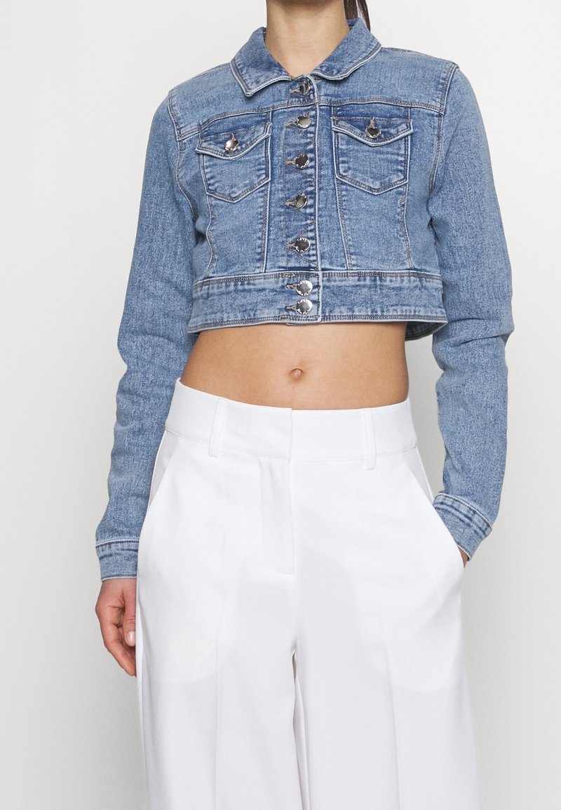 Žena nosící modrou džínovou krátkou bundu se stříbrnými knoflíky a vysokými bílými kalhotami, ruce v kapsách, viditelný trup.