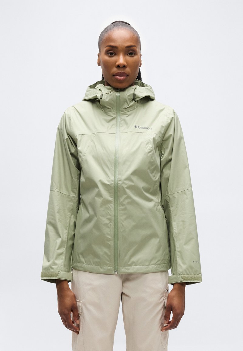 Γυναίκα που φοράει ανοιχτό πράσινο, αδιάβροχο μπουφάν με κουκούλα και φερμουάρ, σε συνδυασμό με ανοιχτό μπεζ παντελόνι, στέκεται μπροστά σε ένα απλό φόντο.